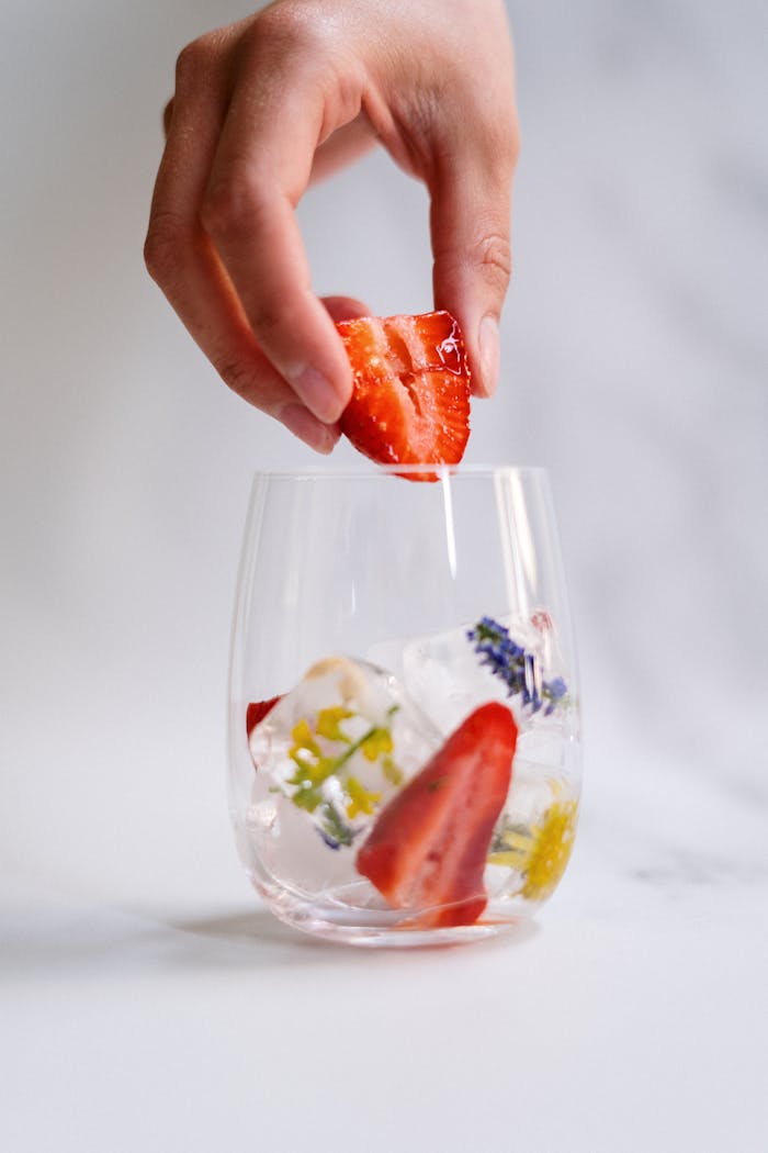 Close-up of a hand adding strawberry to a glass with ice cubes and flower petals.